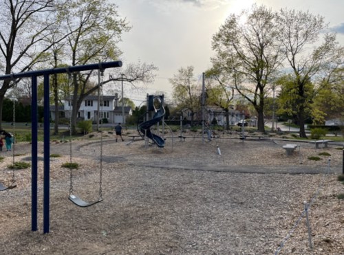 A playground with swings, a slide, and climbing structures, surrounded by trees and houses under a cloudy sky.