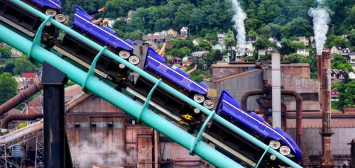 A roller coaster climbs a steep track with a factory and green hills in the background, creating a vibrant scene.