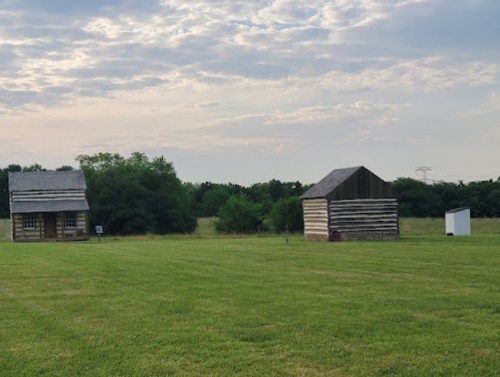 Two historic wooden buildings sit on a grassy field under a cloudy sky, surrounded by trees.