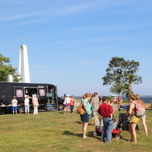 A crowd gathers near a food truck by the water, with a monument and trees in the background on a sunny day.