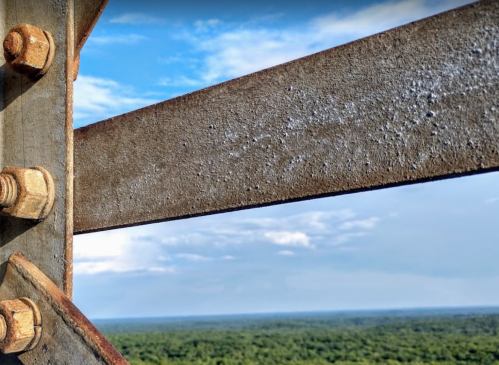 Close-up of rusty metal beams with a view of green treetops and a cloudy sky in the background.