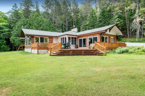 A spacious wooden house with a large deck, surrounded by lush greenery and trees in a serene outdoor setting.