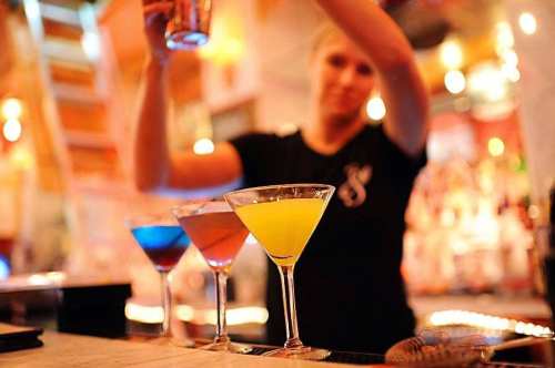 A bartender prepares colorful cocktails at a lively bar, with three drinks in front: blue, red, and yellow.
