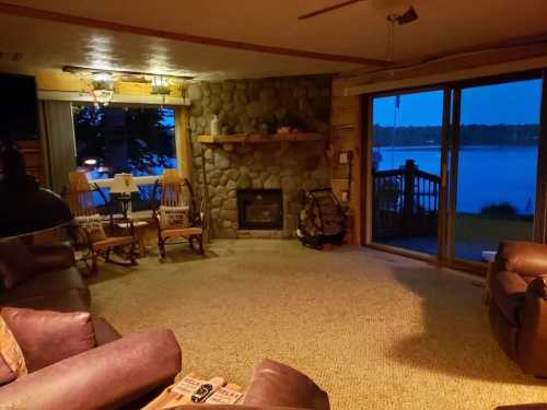 Cozy living room with stone fireplace, rocking chairs, and large windows overlooking a lake at dusk.