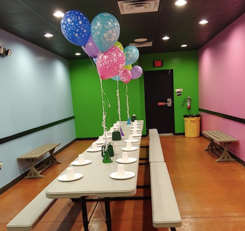 A party room with colorful walls, a long table set with plates, and balloons in various colors floating above.