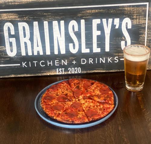 A pepperoni pizza on a blue plate next to a glass of beer, with a sign for Grainsley's Kitchen + Drinks in the background.