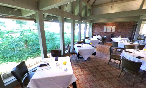 A cozy restaurant interior with tables set for dining, large windows overlooking greenery, and a warm atmosphere.
