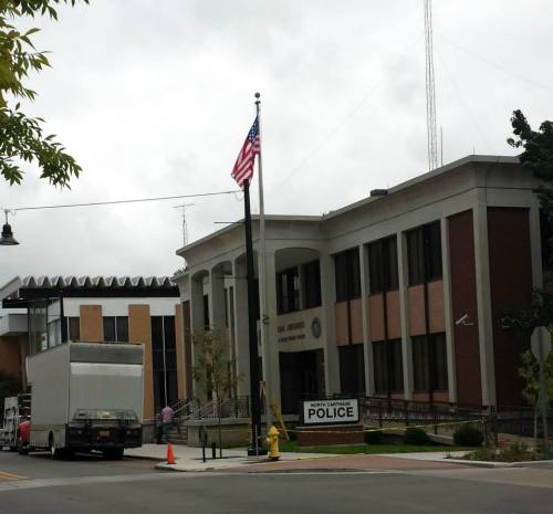 A police station building with an American flag, a moving truck, and a "Police" sign in front. Overcast sky.