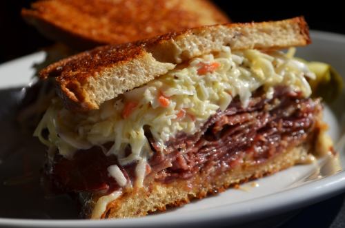 A close-up of a sandwich with layers of meat, coleslaw, and toasted bread, served on a plate.