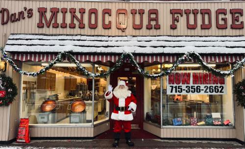 Santa Claus waves outside Dan's Minocqua Fudge shop, decorated for the holidays with snow and festive garlands.