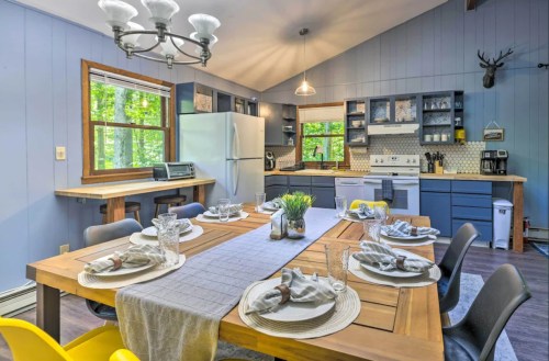 A cozy kitchen with a wooden dining table set for six, modern appliances, and large windows overlooking greenery.