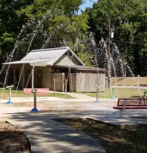 A splash pad with water jets in a park, featuring a small shelter and benches nearby under a clear blue sky.