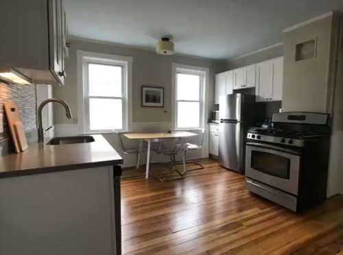 Modern kitchen with stainless steel appliances, wooden floors, and a small dining table by the window.