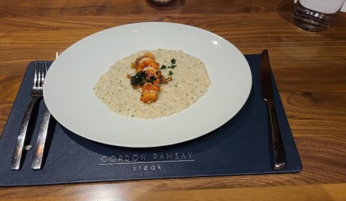 A plate of creamy risotto topped with shrimp and herbs, set on a branded placemat from Gordon Ramsay Steak.