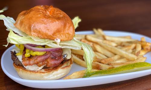 A juicy burger with lettuce, onion, and bacon, served with crispy fries and a pickle on a white plate.