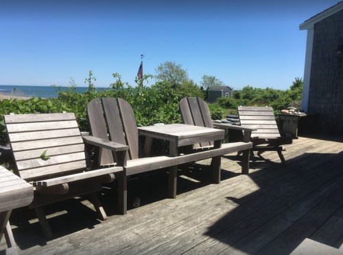 A sunny deck with wooden chairs and a table, overlooking a lush green area and the ocean in the background.