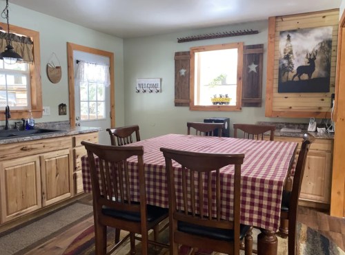 Cozy kitchen with wooden cabinets, a checkered tablecloth, and rustic decor, featuring a large dining table and windows.