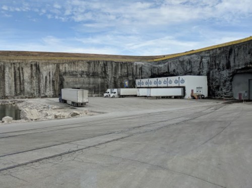 Trucks parked near a large rock wall and a building, with a body of water in the foreground under a blue sky.