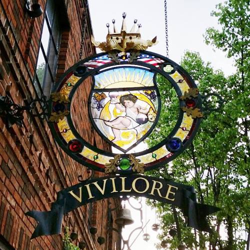 A decorative sign featuring stained glass and ornate details, reading "VIVILORE," surrounded by greenery.