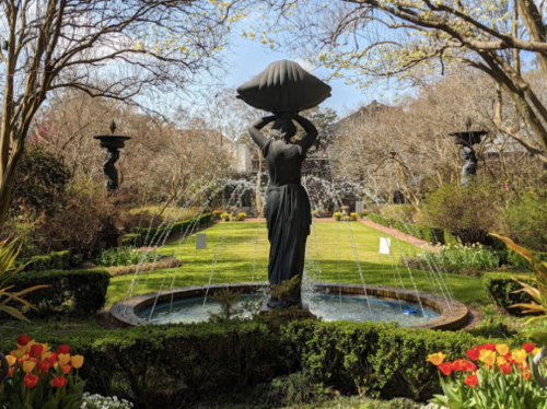 A serene garden featuring a statue holding a shell, surrounded by fountains and colorful tulips in bloom.