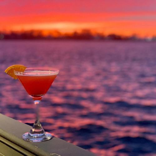 A vibrant sunset reflects on water, with a cocktail glass garnished with an orange slice in the foreground.