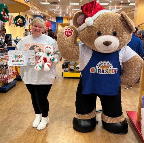 A person stands next to a large teddy bear in a workshop, holding a stuffed animal and a holiday sign.