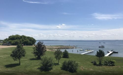 A serene lakeside view with green grass, trees, and a dock, under a clear blue sky with scattered clouds.