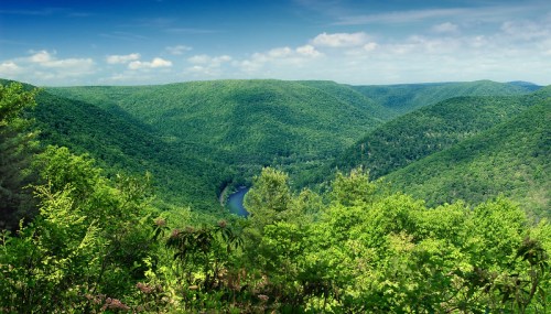 Lush green hills and valleys surround a winding river under a bright blue sky with scattered clouds.