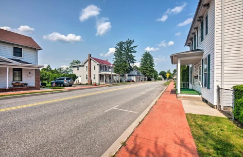 A quiet street lined with houses and trees under a clear blue sky, featuring a brick sidewalk and parked cars.