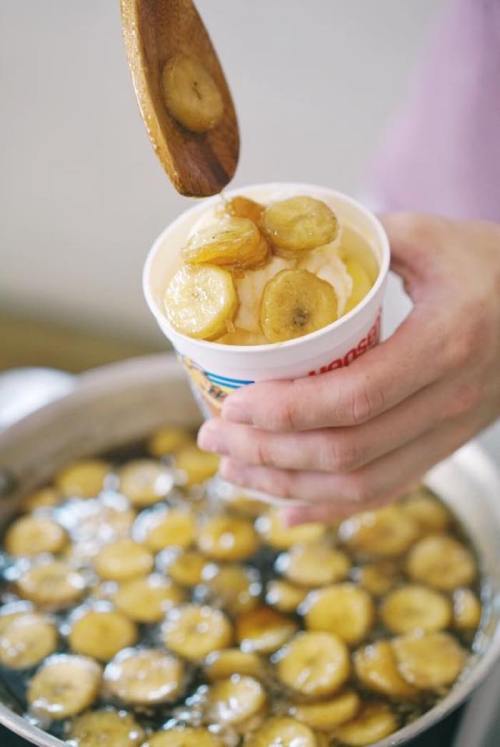 A hand holds a cup of ice cream topped with caramelized bananas, with a pot of fried bananas in the background.