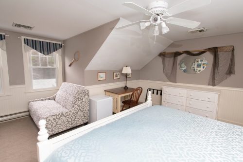 Cozy bedroom with a patterned sofa, desk, lamp, dresser, and ceiling fan under a sloped ceiling.