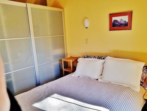 Cozy bedroom with a double bed, yellow walls, and a framed picture above the bed. A wardrobe is visible in the background.