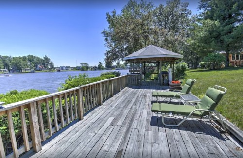 A wooden deck with green lounge chairs overlooks a river and a gazebo surrounded by trees on a sunny day.