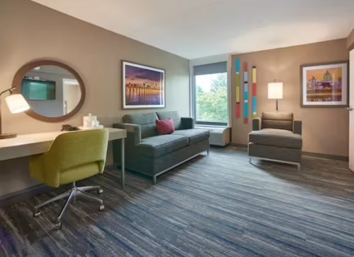 A modern hotel room featuring a desk, sofa, armchair, and large window with colorful artwork on the walls.