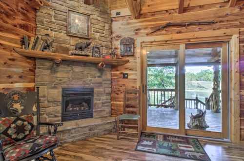 Cozy cabin interior with a stone fireplace, wooden walls, and a view of a deck and nature through glass doors.
