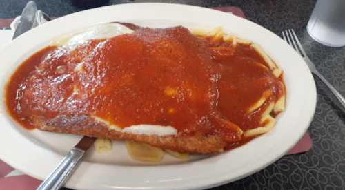 A large plate of chicken parmesan over pasta, topped with marinara sauce, served with a fork on the side.