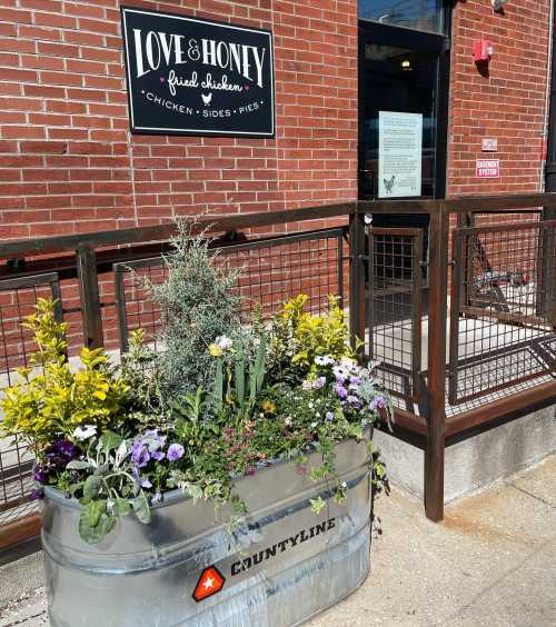 Exterior of Love & Honey Fried Chicken restaurant, featuring a metal planter with colorful flowers and a brick wall backdrop.