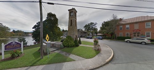 A clock tower in a park near a river, with a sign for Veterans Park and nearby buildings and cars.