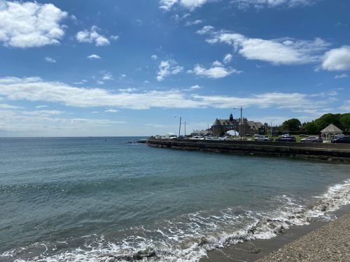 A serene coastal view with gentle waves, a sandy shore, and a building in the background under a partly cloudy sky.