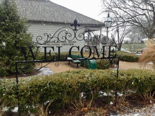 A decorative "WELCOME" sign in a snowy outdoor setting, surrounded by greenery and a building in the background.