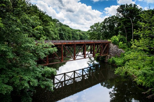 A rusted metal bridge spans a calm river, surrounded by lush green trees and a blue sky with fluffy clouds.