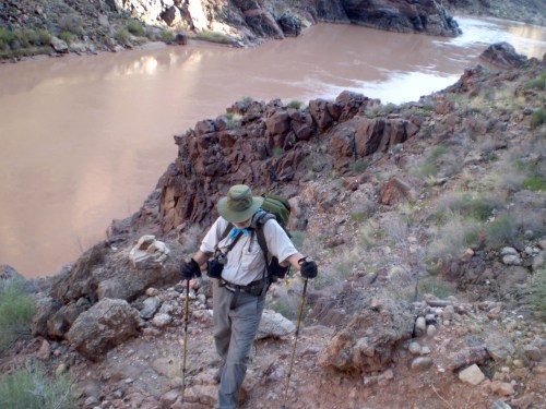 A hiker with a backpack and trekking poles navigates rocky terrain near a river.