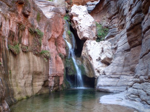 A serene waterfall cascades into a clear pool, surrounded by colorful rock formations and lush greenery.