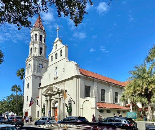 Historic church with a tall bell tower, palm trees, and a clear blue sky, bustling with visitors and parked cars.