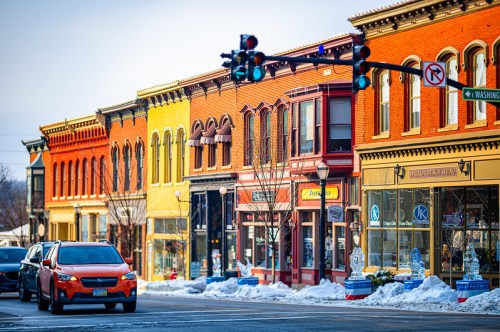 Colorful historic buildings line a snowy street, with a traffic light and parked cars in the foreground.