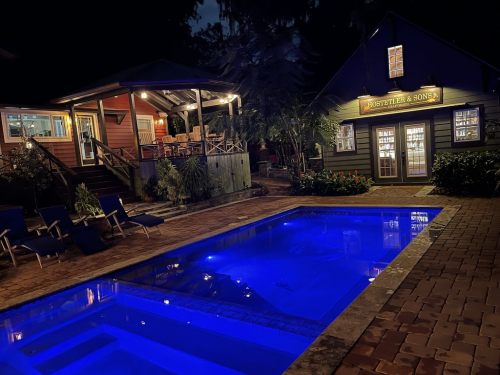 A serene nighttime view of a pool area with blue lighting, surrounded by lush greenery and cozy buildings.