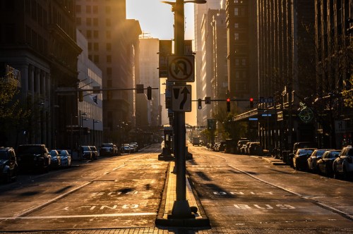 Sunrise casts golden light on a deserted city street, with traffic signals and parked cars lining the road.