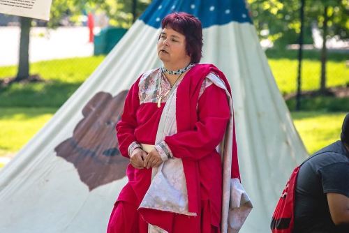 A woman in a red traditional outfit stands near a teepee, looking thoughtfully into the distance.