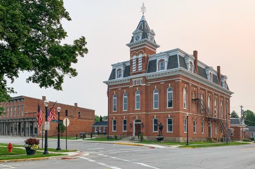 Historic brick building with a clock tower, surrounded by trees and American flags, in a small town setting.