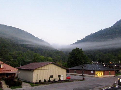 A misty valley between mountains at dawn, with buildings in the foreground and trees lining the hillsides.